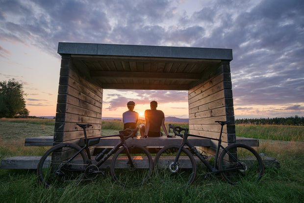 Twee racefietsers zitten in de natuur op een picknick tafel en kijken naar de zonsondergang. Twee CUBE racefietsen staan tegen de picknicktafel aan.