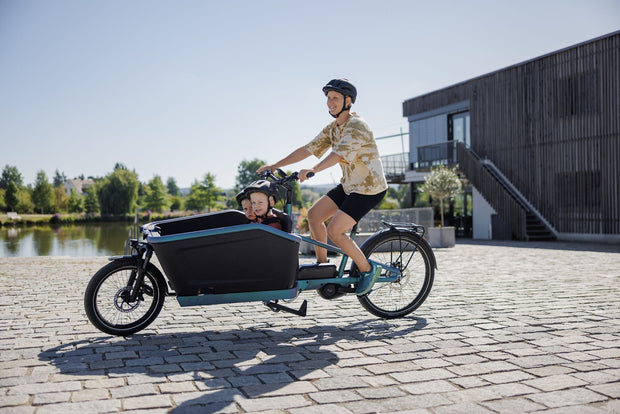 Een moeder rijdt met twee kindjes in een blauwe CUBE Cargo Hybrid- bakfiets. Het is mooi weer en ze rijd op een kade langs het water.. 